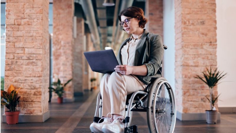 Zu sehen ist eine Frau die im Rollstuhl sitzt und auf ihrem Schoß am Laptop arbeitet. Dies könnte die Dienstunfähigkeit Beamte darstellen.